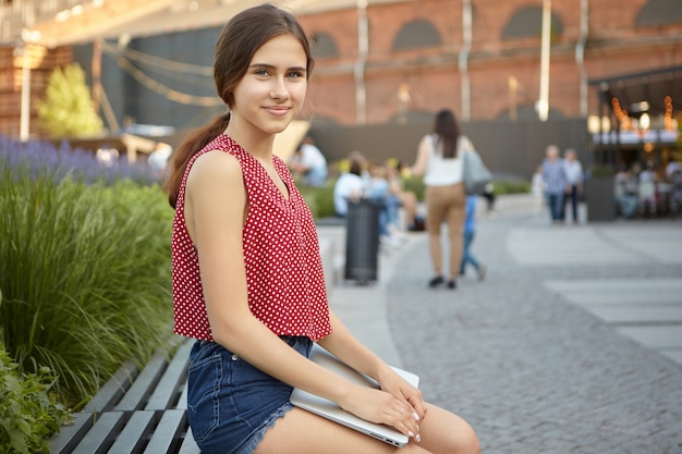 Buiten zomer weergave van mooie positieve jonge vrouw in gestippelde rode top en jeans broek met behulp van draagbaar elektronisch apparaat in park, kijken en glimlachen. Moderne technologie en communicatie