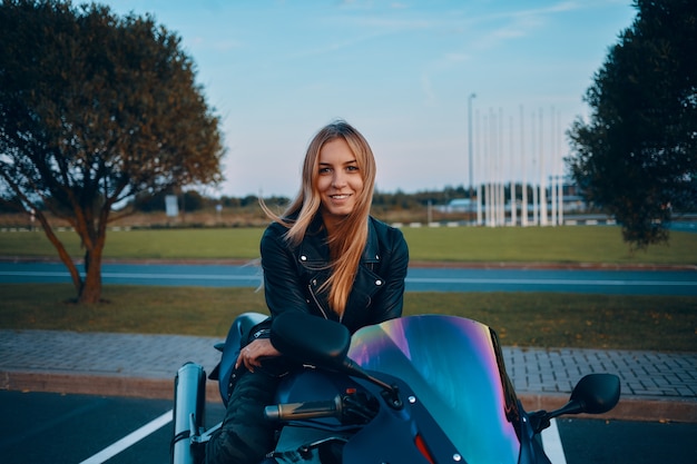 Buitenbeeld van aantrekkelijke jonge Europese vrouw met losse blonde haren, zittend op blauwe motor, gekleed in camouflagejeans en zwart leren jasje