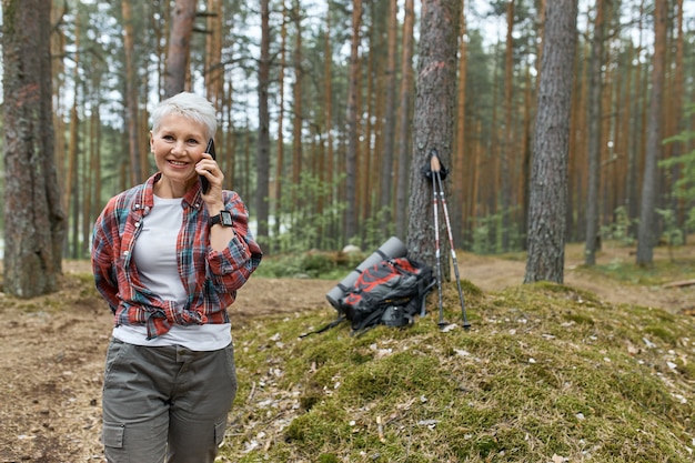 Buitenbeeld van energieke gepensioneerde vrouw in activewear wandelen in het bos, telefoongesprek hebben, glimlachen, rugzak en slaapmat onder boom op achtergrond. Mensen, reizen en technologie