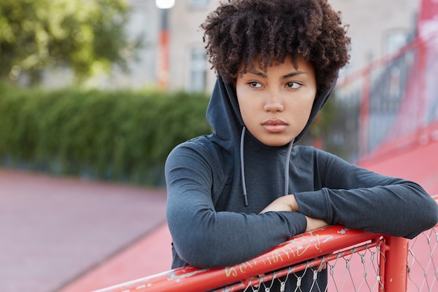 Gratis foto buitenopname van doordachte basketbalspeler in stijlvolle hoodie, poseert op de baan, kijkt peinzend opzij