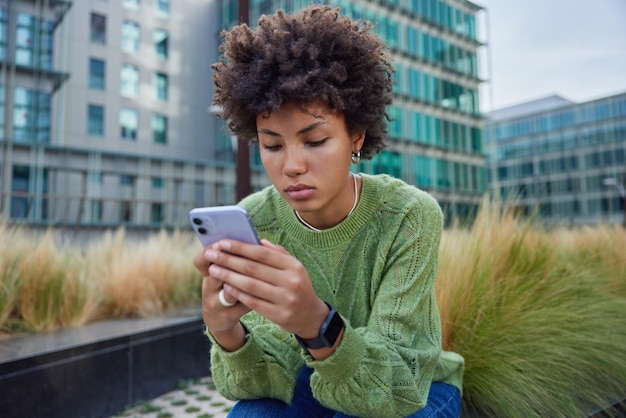 Buitenopname van vrouw met krullend haar gebruikt applicatie op mobiele telefoon voor bankieren gekleed in groene trui zit buiten tegen wazig stedelijke achtergrond overdag creats inhoudspublicatie