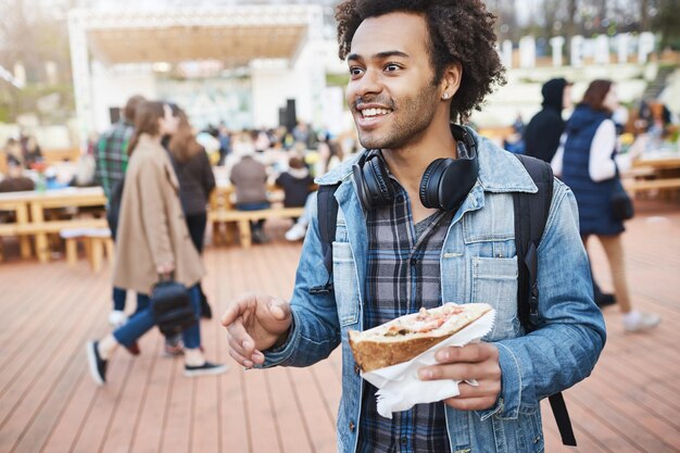 Buitenportret van een positieve Afro-Amerikaanse student die een broodje vasthoudt terwijl hij gebaart en praat met een vriend, op het festival loopt, wachtend op een concert van hun favoriete band.