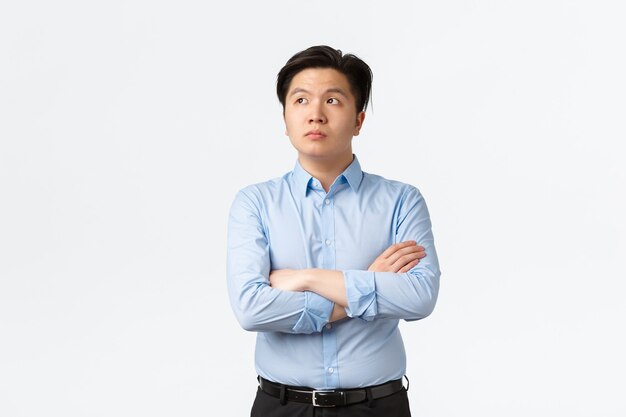 Business, financiën en mensen concept. Doordachte aziatische zakenman in blauw shirt, kruisarmen en kijkend in de linkerbovenhoek, kiezen, denken of dagdromen, staande witte achtergrond