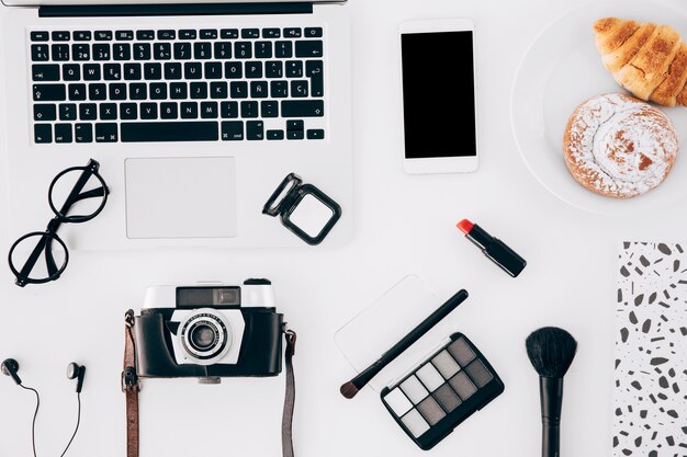 Camera; laptop; mobiele telefoon; cosmetica product en gebakken gebak op witte bureau