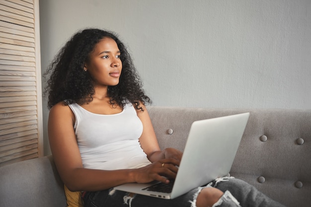 Candid shot van schattige stijlvolle mulat student meisje huiswerk met behulp van laptopcomputer, binnenshuis zittend op een comfortabele bank, peinzende gelaatsuitdrukking. Mensen, technologie en onderwijs