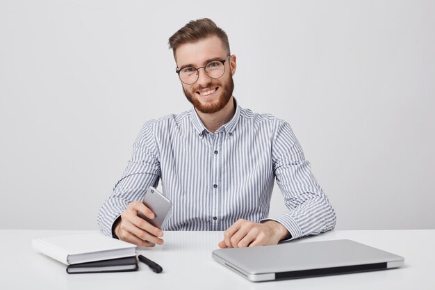 Candid shot van succesvolle man met dikke baard, maakt gebruik van moderne technologie voor werk