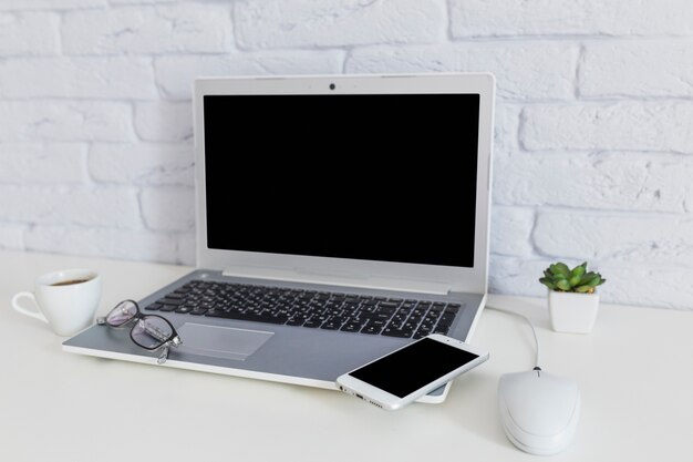 Cellphone en oogglazen op open laptop met koffiekopje over het witte bureau