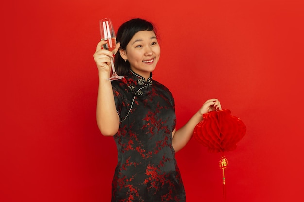 Champagne drinken en lantaarn vasthouden. Gelukkig Chinees nieuwjaar. Aziatisch jong meisje portret op rode achtergrond. Vrouwelijk model in traditionele kleding ziet er gelukkig uit. Copyspace.