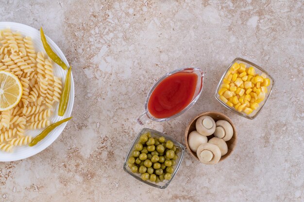 Champignons, maïskorrels, erwten en ketchup in porties naast een schaal pasta op een marmeren oppervlak