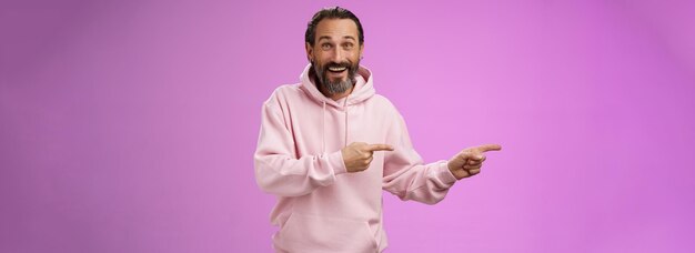 Charismatische, gelukkige, knappe, volwassen man met een stijlvolle hipster hoodie, glimlachend, opgewonden, wijzend.