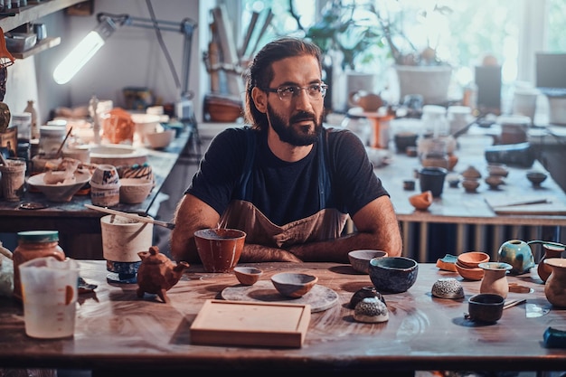 Charismatische gelukkige man met een bril zit in zijn atelier en zijn projecten zijn om hem heen.