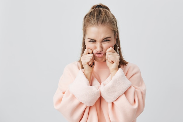 Charmant, fronsend haar gezicht, stijlvol Europees meisje met blond haar gekleed in roze sweatshirt, knijpen haar wangen, spottend, goed humeur en plezier. Plezier, jeugd en schoonheid concept