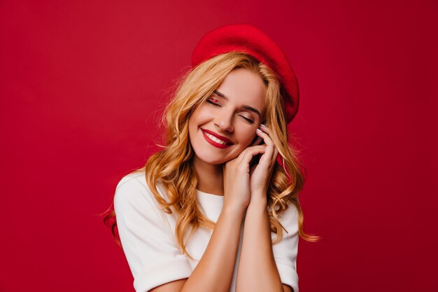 Charmant Kaukasisch meisje dat met golvend kapsel op rode muur lacht. vrolijk Frans vrouwelijk model in baret.