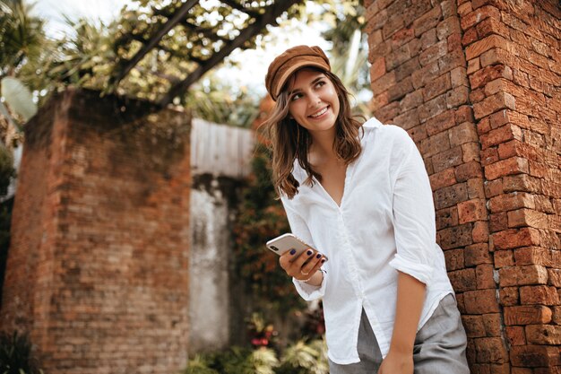 Charmant kortharig meisje leunde tegen de muur van een oud bakstenen gebouw, glimlachend en telefoon vast te houden. Momentopname van vrouw in grijze broek en witte blouse.