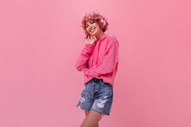 Charmant krullend meisje in denim broek en roze hoodie glimlacht wijd Portret van gelukkige vrouw in koptelefoon luisteren naar muziek op geïsoleerde achtergrond