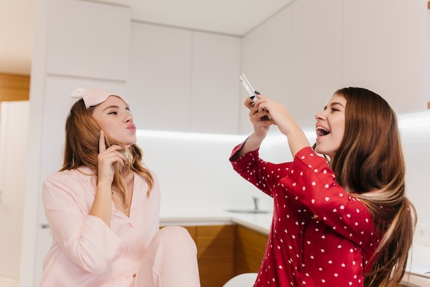 Charmant krullend meisje poseren met kussende gezichtsuitdrukking. Europese vrouw lachen in rood shirt met telefoon voor foto van haar mooie zus.