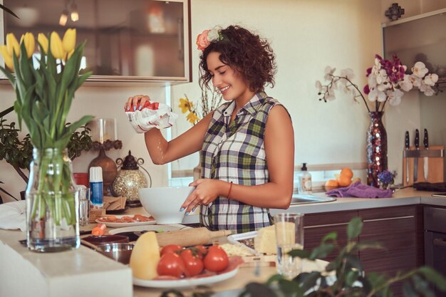Gratis foto charmant krullend spaans meisje giet de bloem in een bord tijdens het koken in haar keuken.