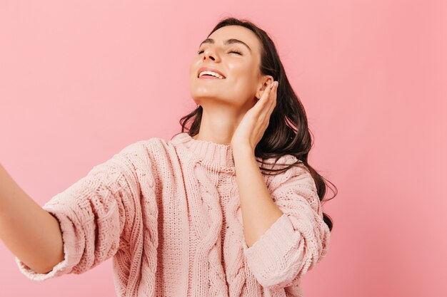 Charmant meisje glimlacht met gesloten ogen. Dame in mooie trui neemt selfie in roze studio.