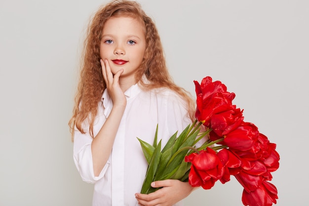 Charmant mooi kind meisje gekleed in witte kleding krijgt een groot boeket rode tulpen als cadeau, kijkend naar de voorkant met zelfverzekerde uitdrukking