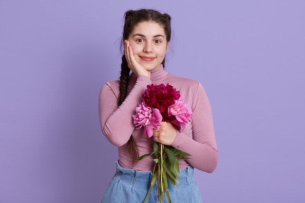 Charmant, mooi meisje met boeket pioenrozen in handen, poseren tegen lila muur, charmante dame met handpalm op wang, vrouw met prettige uitstraling en twee staartjes.