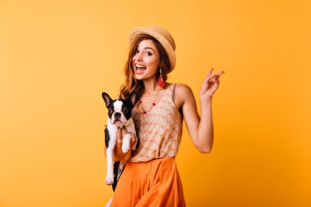 Charmant roodharig meisje in vintage hoed met Franse bulldog. Binnenportret van het verbaasde witte vrouw stellen op geel met puppy.