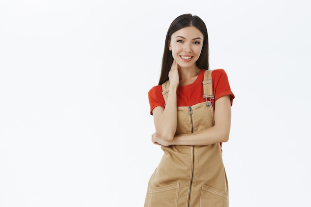 Charmant teder Aziatisch meisje in bruine overall over rood t-shirt raakt nek zachtjes verlegen en onhandig