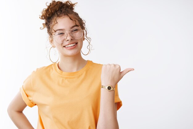 Charmant, vriendelijk ogend schattig gembermeisje rommelig krullend haarknot met oranje t-shirt bril wijzend duim naar rechts glimlachend vragen vriend wil iets voorstellen plaats zitten samen koffie drinken