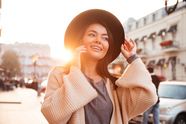 Charmante Aziatische vrouw die haar zwarte hoed aanpast terwijl het spreken op mobiele telefoon op stadsstraat