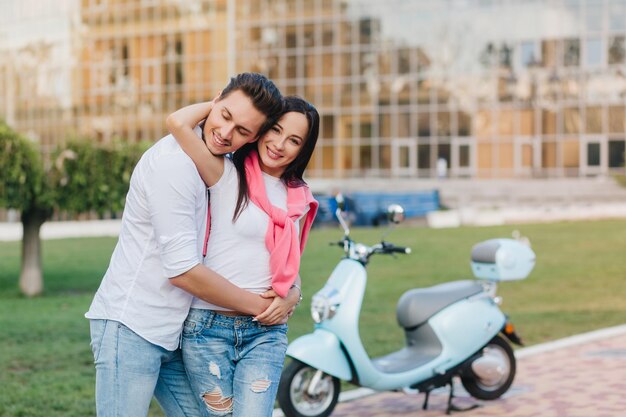 Charmante blanke vrouw met roze trui die echtgenoot omhelst tijdens fotoshoot in park. Gelukkig getrouwd jong stel poseren naast scooter op de achtergrond van de stad wazig.