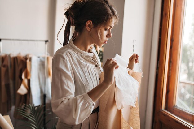 Charmante dame die naar haar ontworpen kleding kijkt Aantrekkelijke jonge vrouw in witte blouse werkt aan haar nieuwe modecollectie