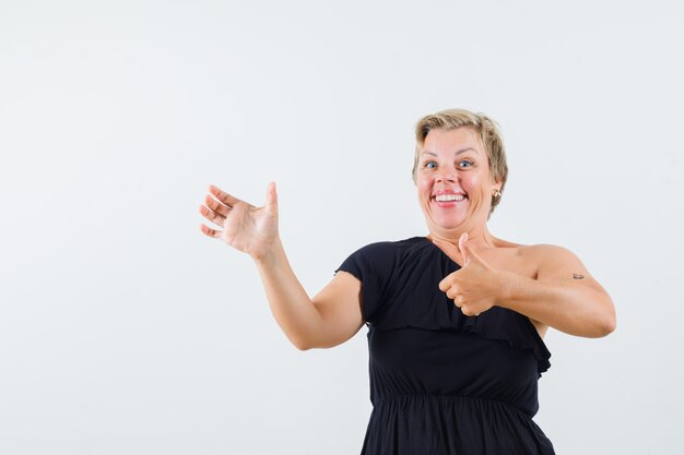 Charmante dame die zich voordeed als het vasthouden van de telefoon terwijl de duim in een zwarte blouse wordt weergegeven en er vrolijk uitziet. vooraanzicht.