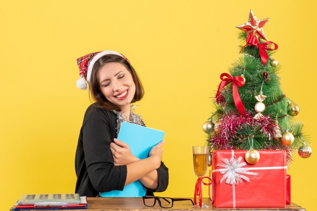 Charmante dame in pak met kerstman hoed en nieuwjaarsversieringen document omarmen en dromen in het kantoor op geel geïsoleerd