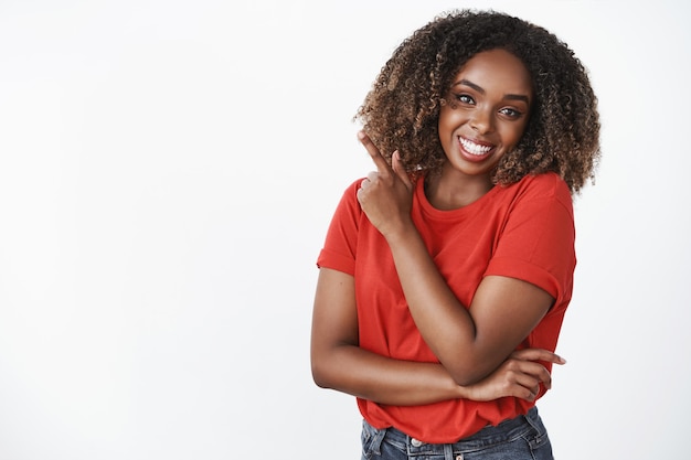 Charmante flirterige en vrouwelijke zorgeloze Afro-Amerikaanse jonge vrouw met krullend haar in rood t-shirt glimlachend vreugdevol wijzend op de linker bovenhoek over witte muur