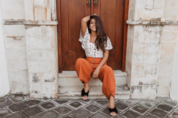 Charmante gekrulde brunette vrouw in culottes en witte blouse zit op drempel van huis met houten deur