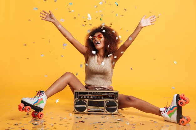 Charmante jonge Afrikaanse vrouw in retro slijtage en rolschaatsen die confettien werpen terwijl het zitten met boombox