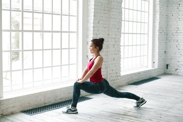 Charmante jonge blanke vrouwelijke yoga-instructeur met haarknoop die benen opwarmt voor de les in het sportschoolcentrum, vrolijk glimlachend tijdens het doen van een hoge lunge pose. Leuk meisje dat overdag binnenshuis uitoefent