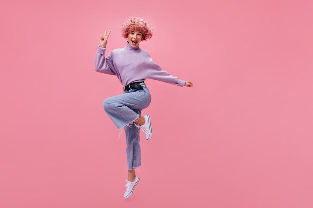 Charmante jonge vrouw in denim broek en paarse trui springt op roze achtergrond Vrolijk krullend roze meisje in jeans glimlacht en toont vredesteken op geïsoleerd