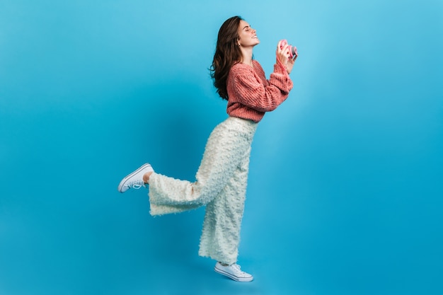 Charmante jonge vrouw in witte broek maakt foto op roze camera. Portret van gemiddelde lengte van brunette in roze sweater.