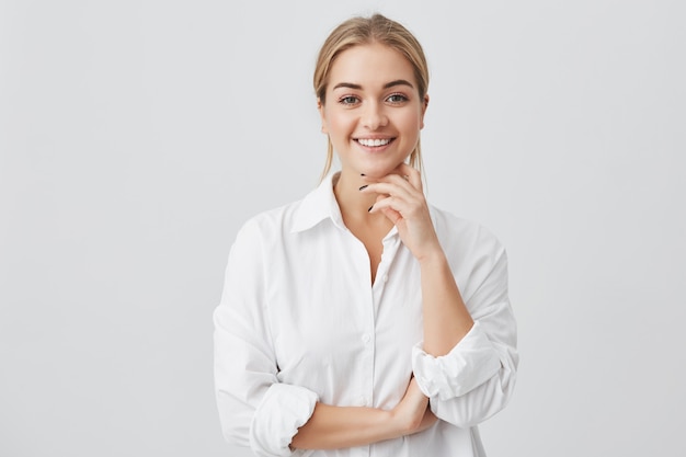 Charmante jonge vrouw met blond steil haar, gekleed in een wit overhemd dat aangenaam in de camera kijkt en haar kin aanraakt. Jong mooi meisje met het brede glimlach stellen.