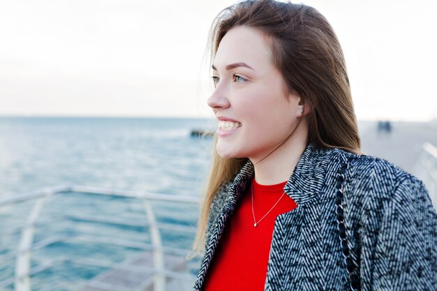 Charmante, lieftallige vrouw met lang lichtbruin haar in een grijze vacht om de zee en het mooie weer op de kade te bewonderen