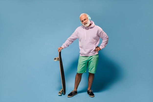 Charmante man in hoodie en groene korte broek die naar muziek luistert en een skateboard vasthoudt