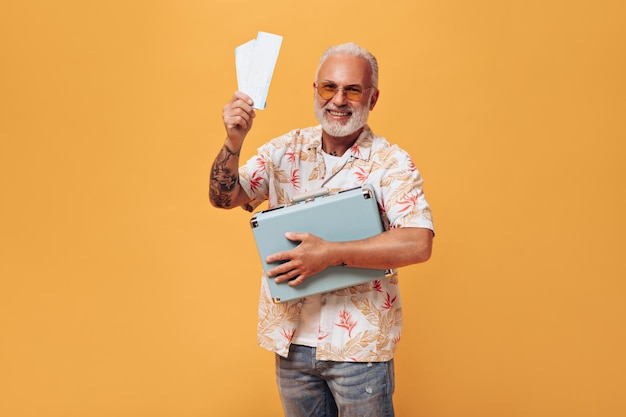 Charmante man in plant print shirt poseert met kaartjes en koffer op oranje achtergrond Grijsharige man met baard in zomerkleren heeft plezier