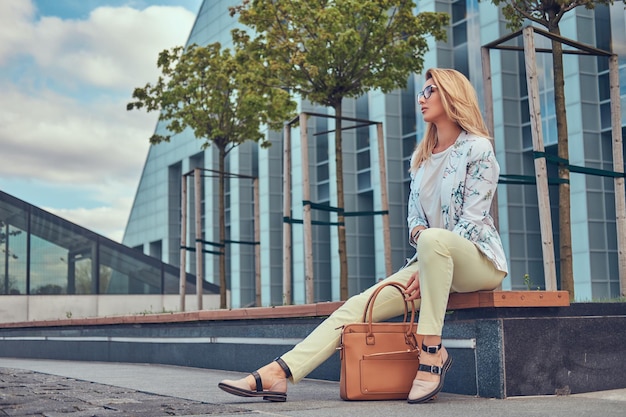 Charmante modieuze vrouw in stijlvolle kleding en bril met een handtas, zittend op een bankje tegen een wolkenkrabber.