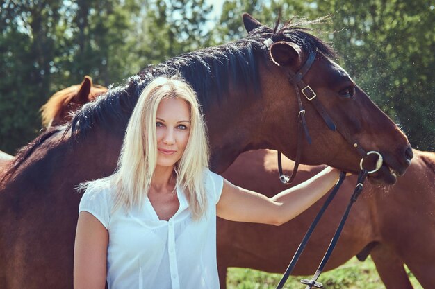 Charmante mooie blonde, gekleed in een witte blouse en spijkerbroek, knuffelen een paard naar een groen veld.