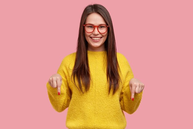 Charmante, onbezorgde vrouw met een positieve uitdrukking, wijst met beide wijsvingers naar beneden, gekleed in vrijetijdskleding, heeft een brede glimlach