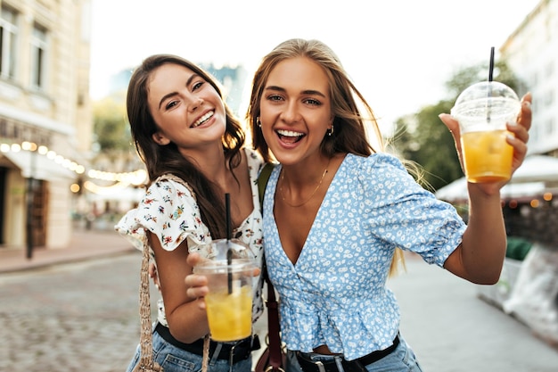 Charmante opgewonden blonde en brunette vrouwen in gebloemde blouses en jeans verheugen zich veel plezier buiten wandelen in het stadscentrum en genieten van limonade