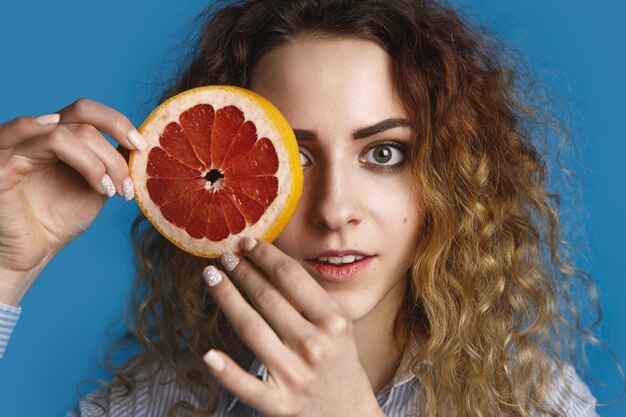 Charmante schattige jonge vrouw met groene ogen en volumineus haar poseren, een oog verbergen achter ronde sappige rijpe grapefruit. Versheid, vitamines, gezondheids-, zorg-, schoonheids- en wellnessconcept