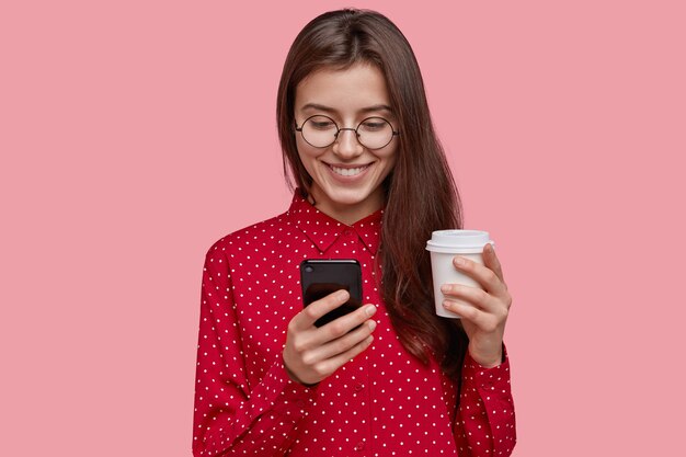 Charmante, tevreden jonge vrouw houdt warme afhaalkoffie, mobiele telefoon, blij om een nieuw apparaat als cadeau te ontvangen, draagt een rood shirt, had een zachte glimlach