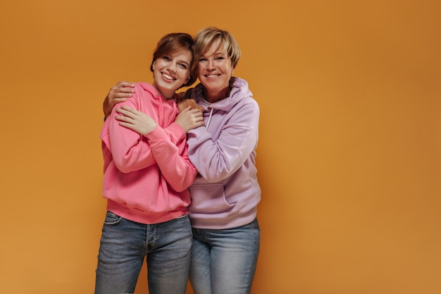 Charmante twee dames met een mooie glimlach en kort cool kapsel in moderne roze sweatshirts en trendy jeans knuffelen op geïsoleerde achtergrond.