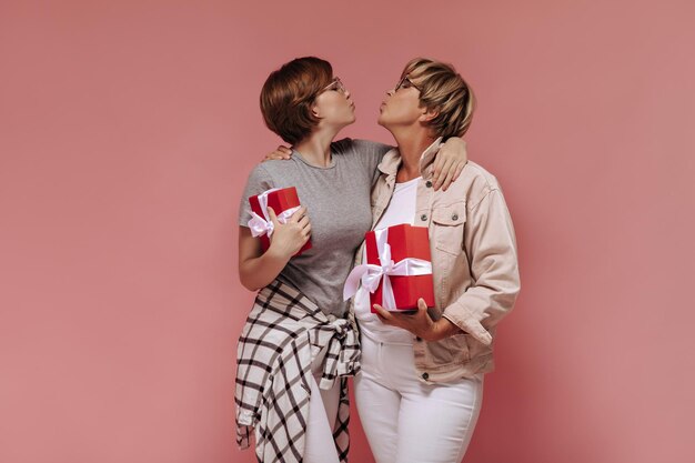 Charmante twee dames met kort cool kapsel in trendy kleding die kus blazen en rode geschenkdozen op roze achtergrond houden